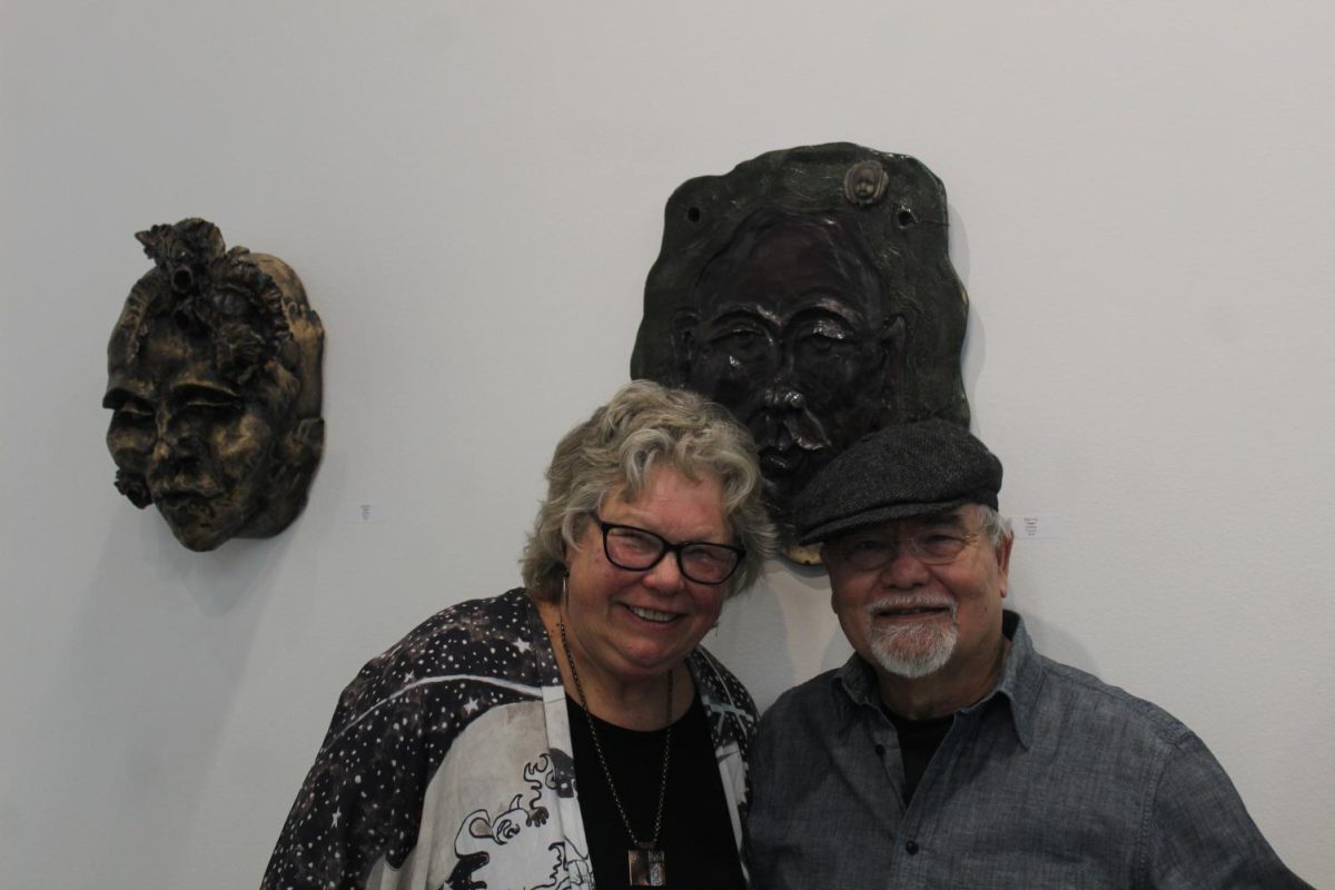 Artists Katri Uno and Roy Lizama pose in front of Uno's clay masks in the Visual and Performing Arts Center. The "Vessel and Visage" art exhibition featured works from both artists.